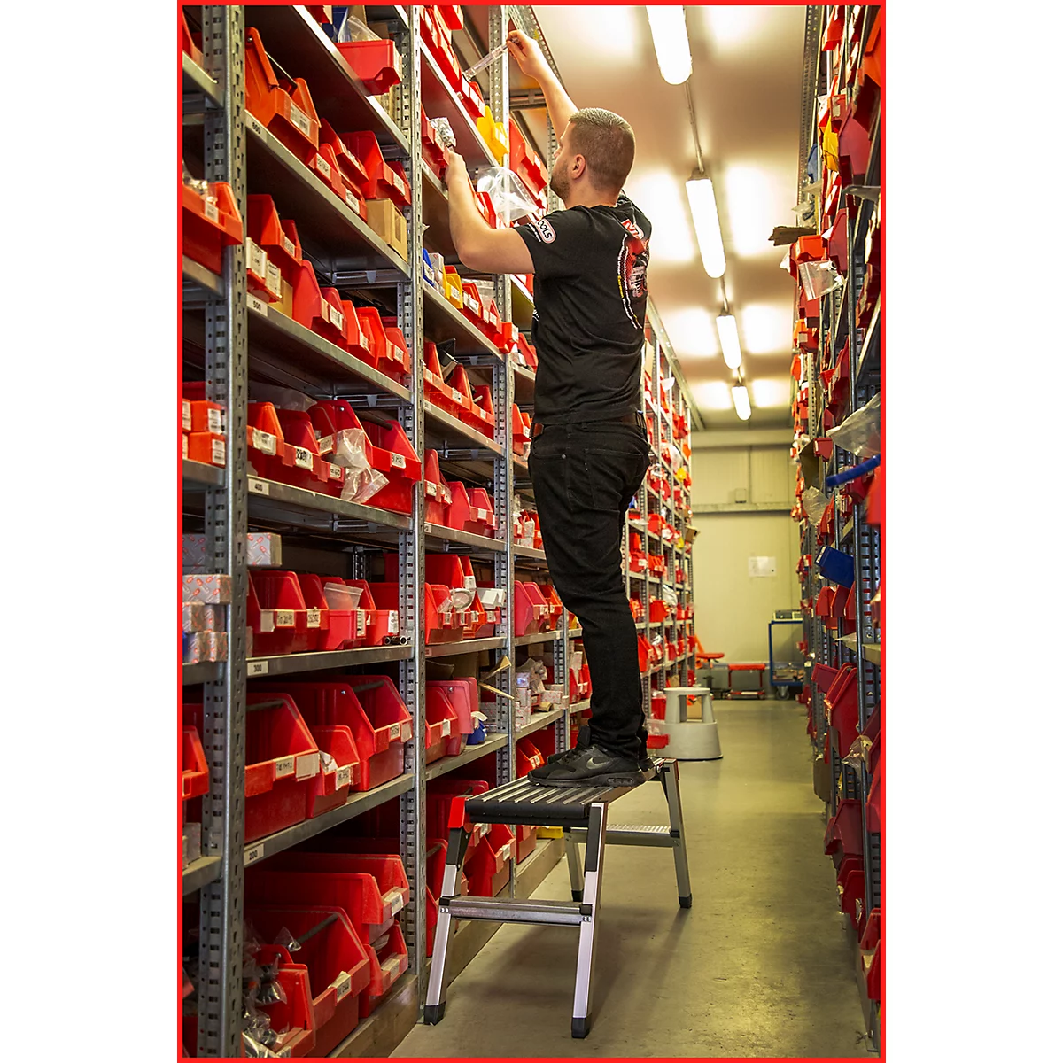 Ein Mann steht auf einer Trittleiter und greift in ein Regal voller roter Behälter. Er trägt ein schwarzes T-Shirt und eine schwarze Hose.